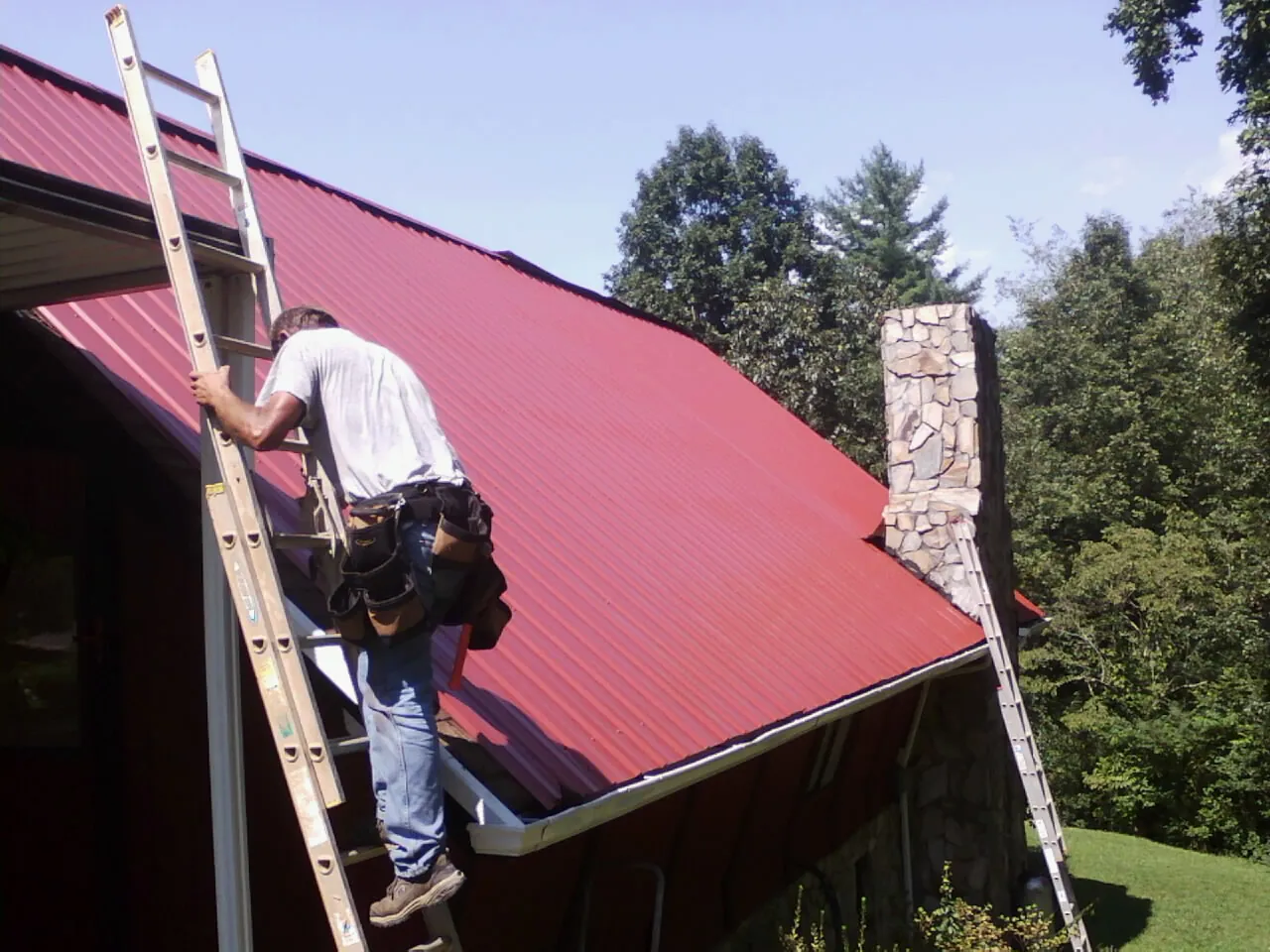 Roofer on a ladder beside a red metal roof and stone chimney — Soffit & Fascia Repair by Roofing Brothers in Upperstrasburg